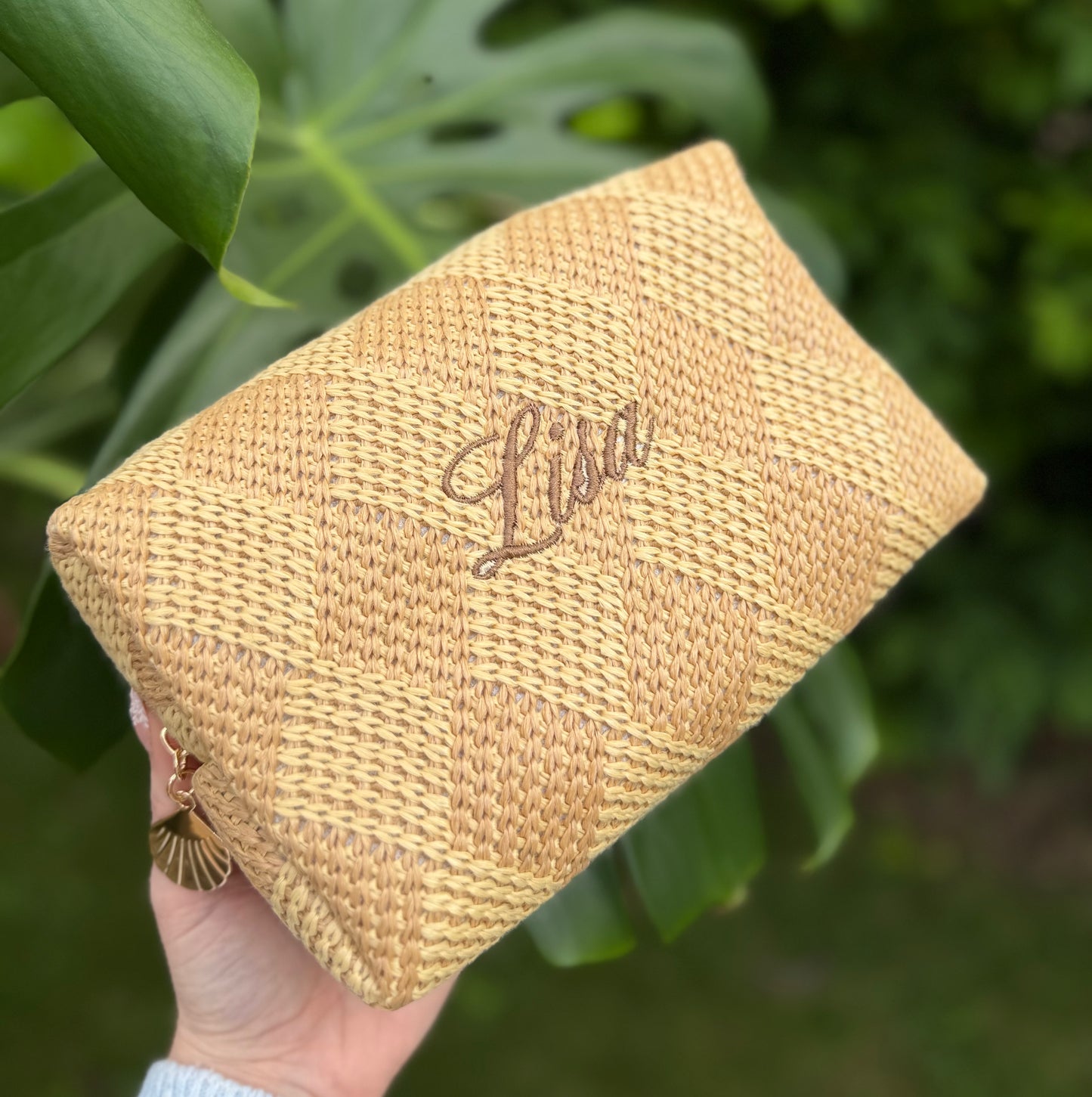 Woven pouch with 'Lisa' embroidery held in a hand against a green leafy background